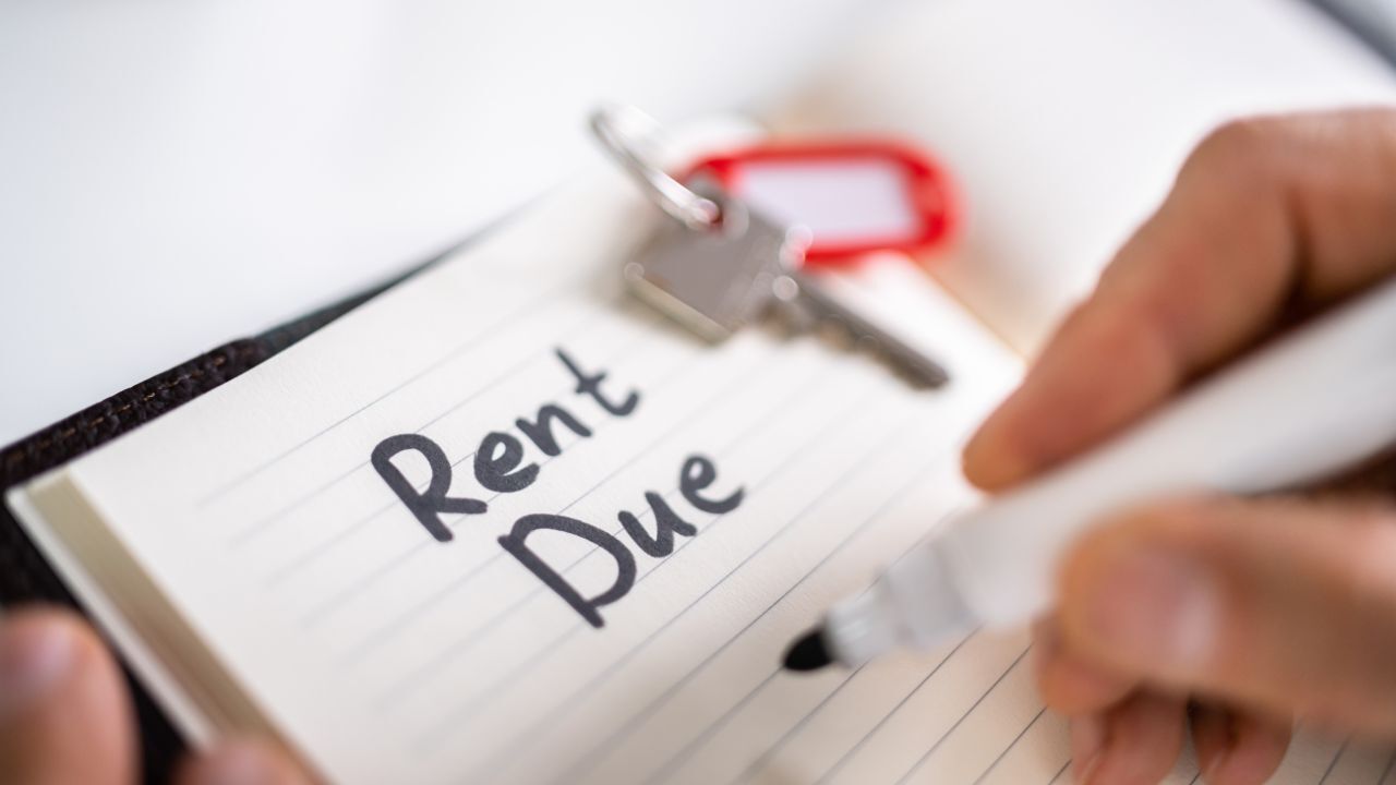 Close-up of a hand writing “Rent Due” in a notebook with a house key placed on top.