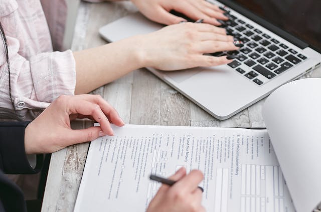 A person filling in a document next to another person using a laptop